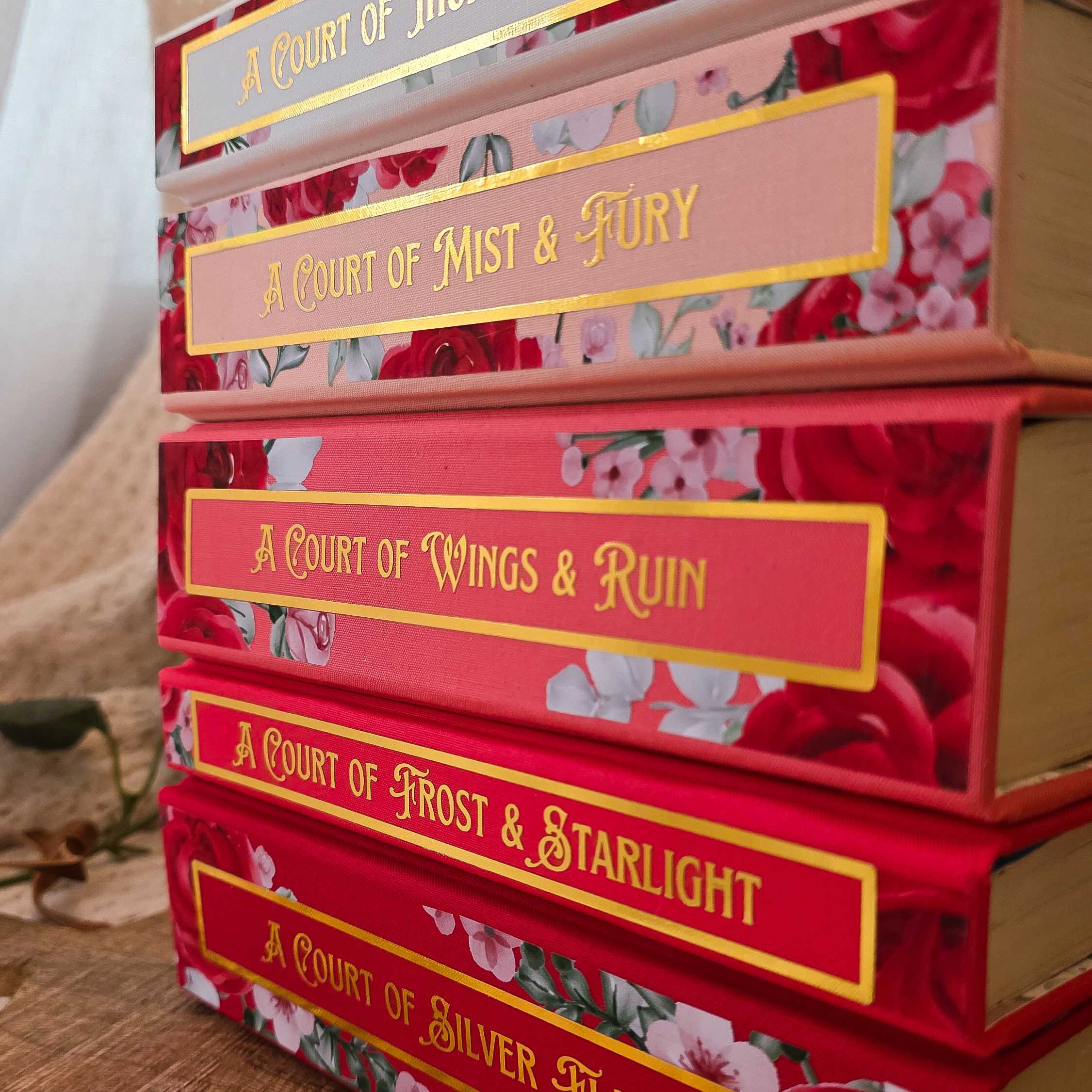 Stack of books titled 'A Court of Mist & Fury', 'A Court of Wings & Ruin', 'A Court of Frost & Starlight', and 'A Court of Silver' with floral designs on the covers.
