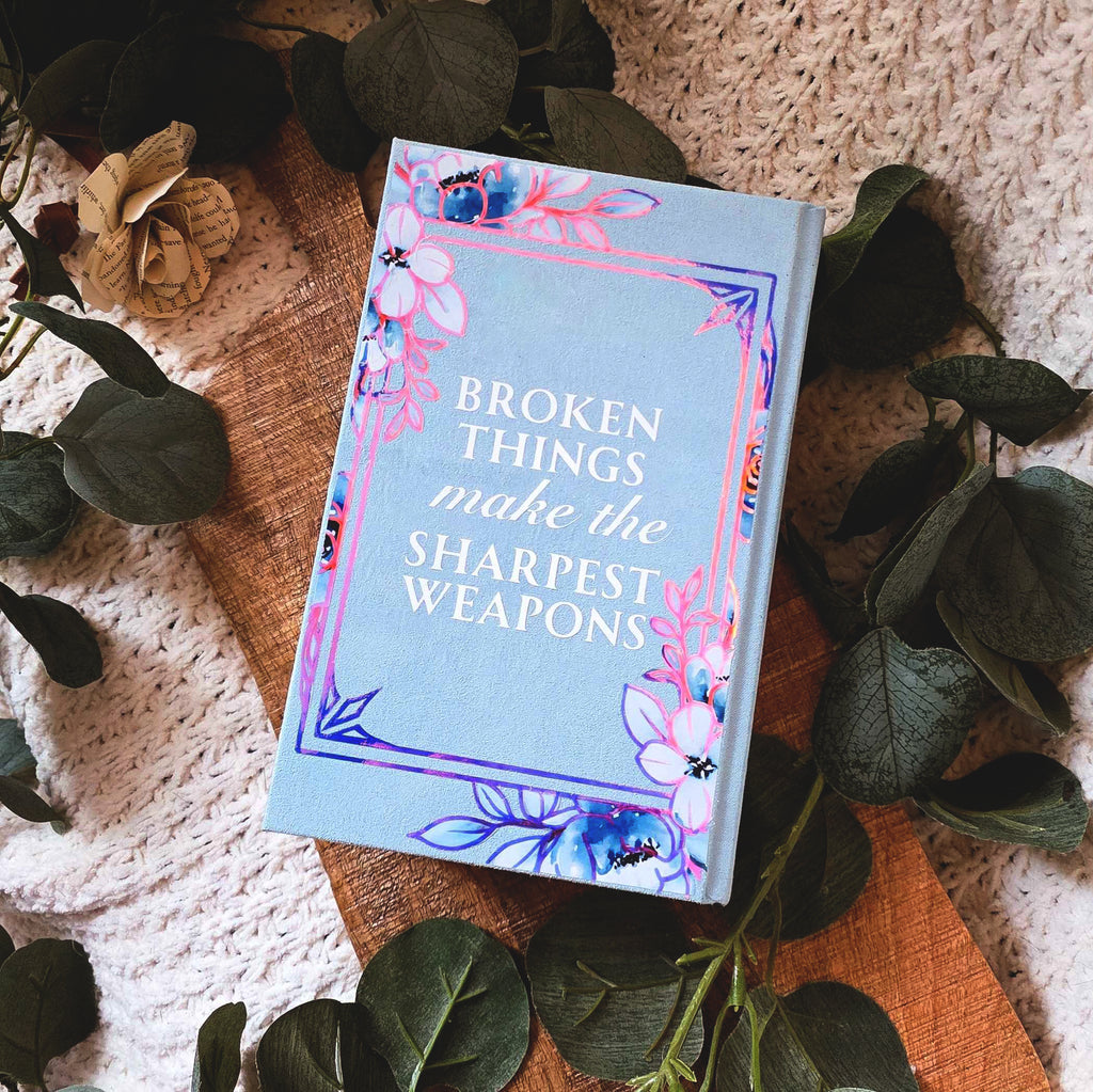 Book quote 'Broken Things Make the Sharpest Weapons' with floral design on a textured surface with leaves.